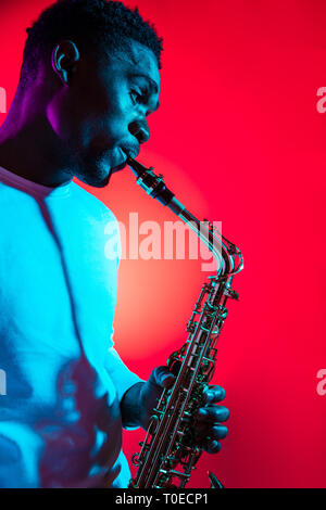 African American handsome jazz musician playing the saxophone in the studio on a neon background. Music concept. Young joyful attractive guy improvising. Close-up retro portrait. Stock Photo