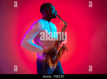 African American handsome jazz musician playing the saxophone in the studio on a neon background. Music concept. Young joyful attractive guy improvising. Close-up retro portrait. Stock Photo