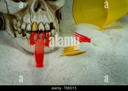Pirate skull toy with red plastic fork in teeth, straws, disposable glass and sand, viewed from high angle in close-up. Funny Jolly Roger, plastic was Stock Photo