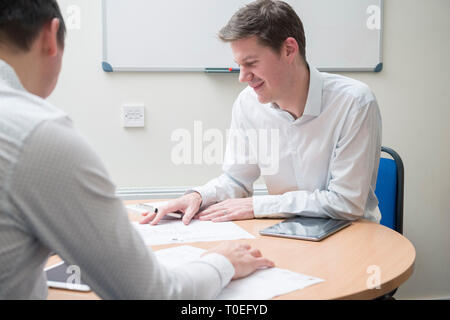 Two men sit round a table in a small office meeting room to review plans in a sales meeting Stock Photo