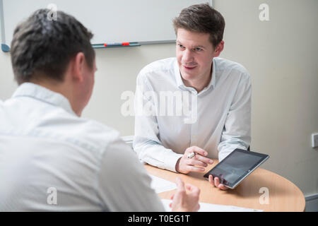 Two men sit round a table in a small office meeting room to review plans in a sales meeting Stock Photo