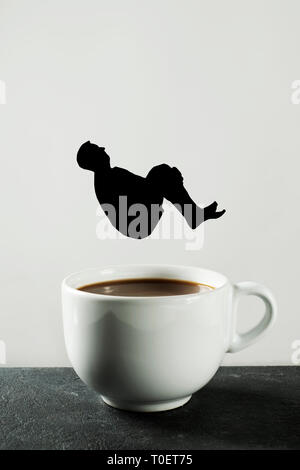 a black paper cutout in the shape of a man throwing himself into a white ceramic cup with coffee, on a dark stone surface against a white background w Stock Photo