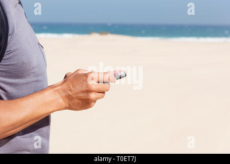 closeup of a young caucasian man, wearing a T-shirt and carrying a backpack, using a smartphone on a white sand beach with the ocean in the background Stock Photo