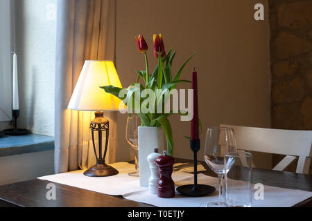 Dining wooden table covered with white tablecloth. Lamp, candles, floral bouquet of red tulips flower in vase and glasses on served table for romantic Stock Photo