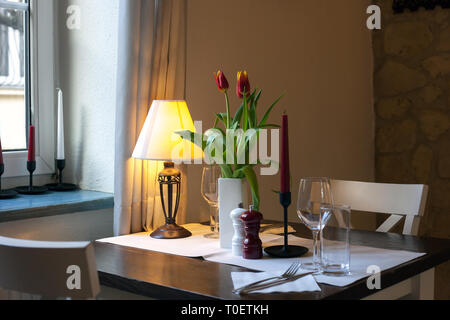 Dining wooden table covered with white tablecloth. Lamp, candles, floral bouquet of red tulips flower in vase and glasses on served table for romantic Stock Photo