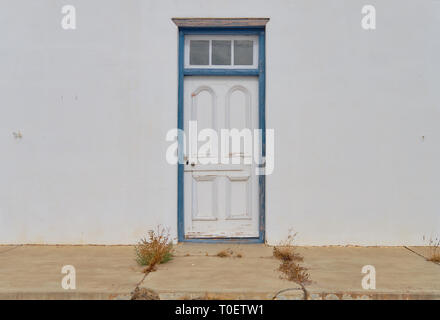 Minimalist capture of a single rural door on a white textured wall.  Door has blue frame in a state of needing repairs. Front concrete porch with weed Stock Photo