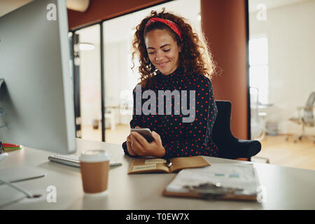 Smiling young businesswoman reading a text message on her cellphone while sitting at her desk in an office Stock Photo