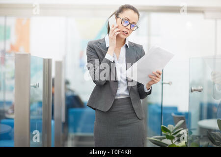 Portrait of successful young businesswoman speaking by phone and using digital tablet while standing in office, copy space Stock Photo