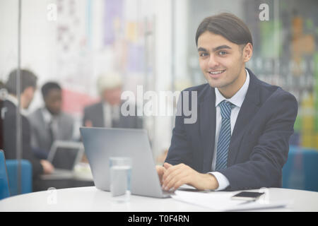Portrait of young Middle-Eastern businessman looking at camera and using laptop while working in office sitting behind glass, copy space Stock Photo