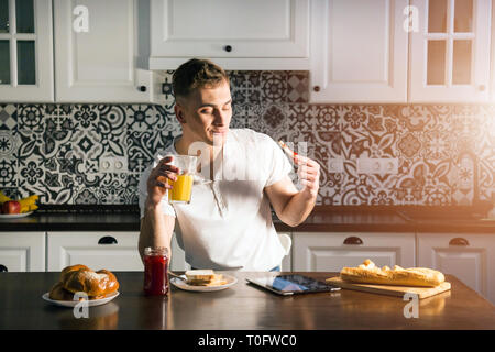 Guy eating tosts with strawberry and drinking juice while using tablet at the morning kitchen Stock Photo
