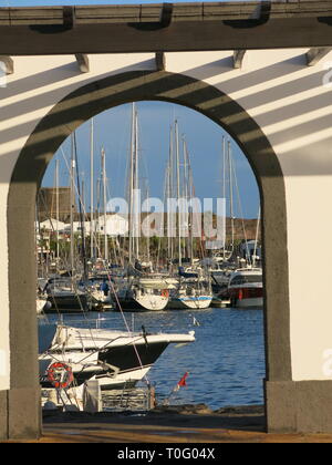The evening sun casts shadows on the wall above the view through a stone archway of the boats moored up in the harbour at Rubicon Marina, Playa Blanca Stock Photo