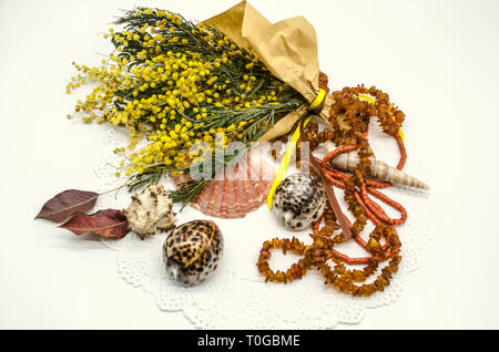 Still life with a bouquet of yellow mimosas, wrapped in rough paper and tied with ribbons, with seashells, amber beads and coral beads on a delicate w Stock Photo