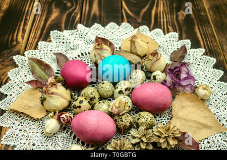 Knitted openwork white napkin with Easter eggs and quail eggs among dry rose buds and dry leaves on dark polished boards Stock Photo