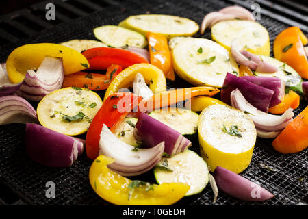 Fresh vegetables, including peppers, red onion, zucchini, and yellow squash, with olive oil and fresh herbs, roasting on a grill with cooking mesh. Stock Photo