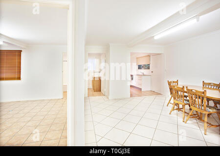 Bright white clean home interior with minimalist decor and a view down a dividing wall to an empty tiled room and dining area with table and chairs Stock Photo