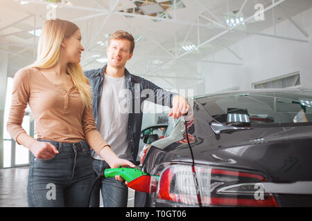 Young smiling family couple buying first electric car in the showroom. Close-up of attractive woman charging ecological hybrid car with the power Stock Photo