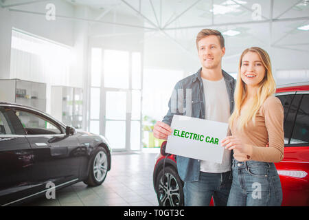 Young family buying first electric car in the showroom. Smiling attractive couple holding paper with word Electric car while standing near eco red Stock Photo