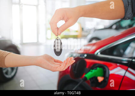 Young family buying first electric car in the showroom. Close-up of male hand giving car key to female hand on battery electric car background. Eco Stock Photo