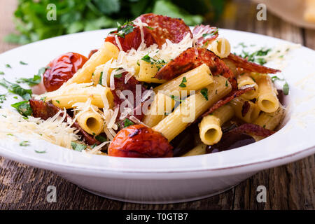 Rigatoni pasta with salami, roasted cherry tomatoes, and olives, delicious Italian dish served with grated Parmesan cheese on rustic wooden table Stock Photo
