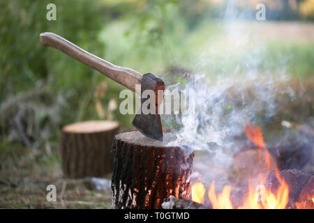 Axe in tree stump and campfire with smoke in summer forest at evening Stock Photo