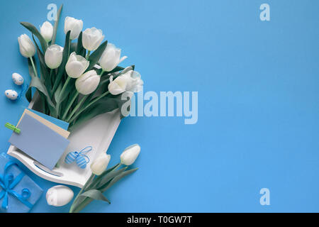 Easter border on blue paper background with copy-space. White tulips, colored eggs, wrapped gift and blank greeting cards on white tray, toned image. Stock Photo