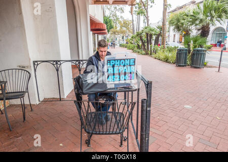 An iPhone repairman sets up shop on the sidewalk patio of a local coffee shop with a homemade sign stating "iPhone Repair" and his phone number. He si Stock Photo