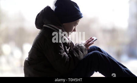 boy uses the phone, sits on the window frame of the old building in winter Stock Photo