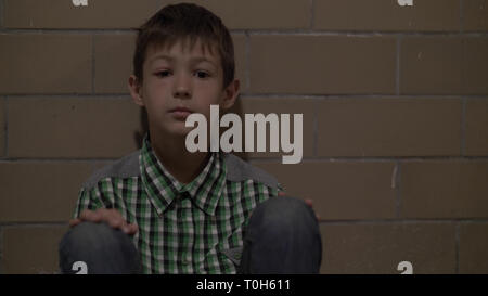 sad lonely battered boy sits on the floor lokking at the camera, no one is waiting for the boy at home Stock Photo