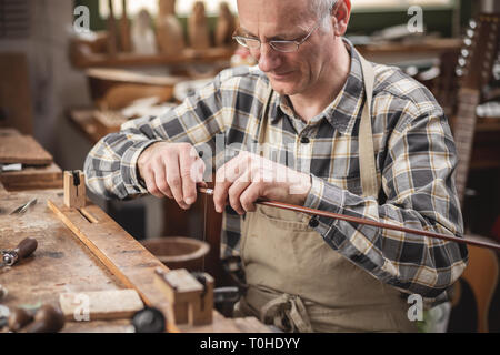 Mature instrument maker inside a rustic workshop is winding a wire around a violin bow