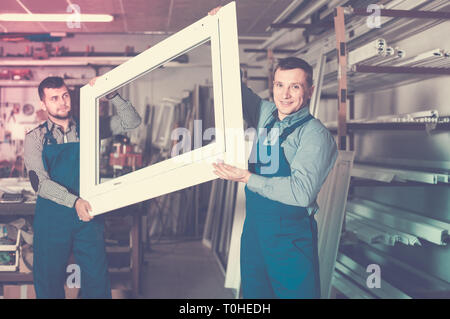 Production smiling workers in coverall with different    finished PVC profiles and windows at factory Stock Photo