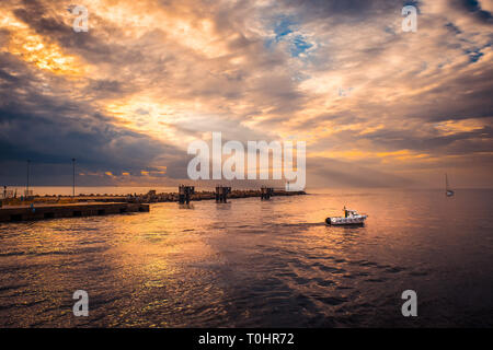A fishing boat returns to the harbor at home by the sea at sunset lit by the rays of the sun, after a day of work Stock Photo