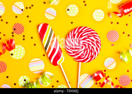 Two multicolored candies among paper circles of confetti, glitter and festive ribbons on a bright yellow background. Top view, flat lay Stock Photo