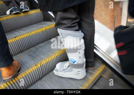 Mans lower leg wearing a plastic leg protection splint or walking cast to secure his leg that has a medical problem. He is standing on the moving escu Stock Photo