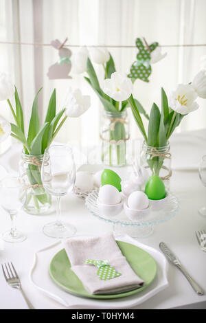 Decor and table setting of the Easter table with white tulips and dishes of green and white color. Easter decor in the form of Easter bunnies  green c Stock Photo