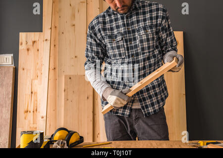 Carpenter is holding small pine plank in workshop over his working desk with tools for woodwork crafts Stock Photo