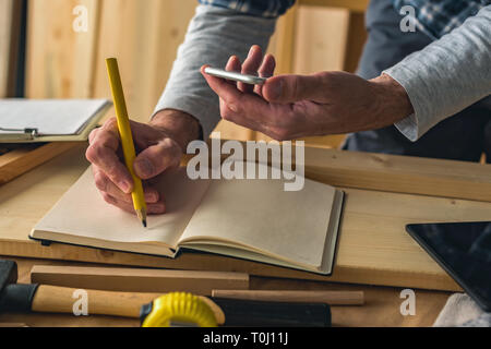 Carpenter is writing project notes while using smartphone in small business woodwork workshop Stock Photo
