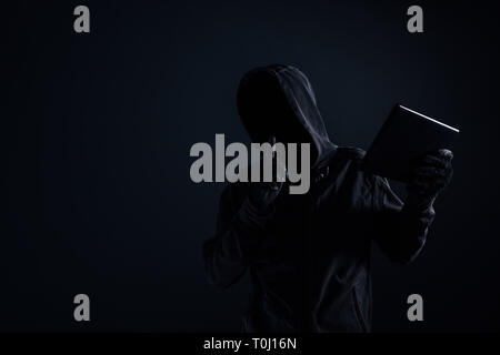 Hooded computer hacker with obscured face using digital tablet in cybercrime and cybersecurity concept, low key with selective focus Stock Photo