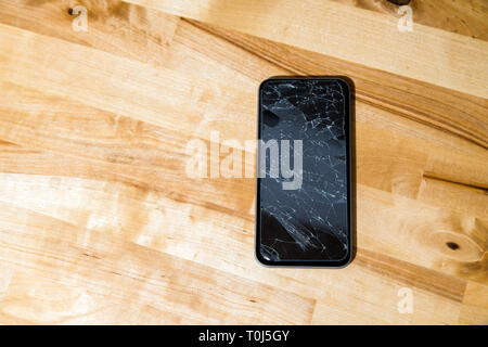 Concept of smart phone with broken screen. Top view on wooden desk background. Cracked, shattered lcd touch screen on modern cellphone. Gadget needs r Stock Photo