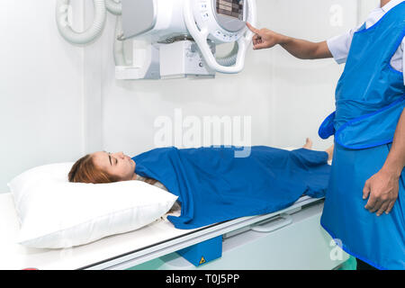 Asian young woman patient lying under X-ray device with radiologist preparing machine in X-ray room at hospital. Stock Photo