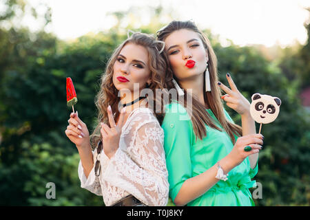 Two pretty and fashionable girlfriends holding candies on stick. Stock Photo