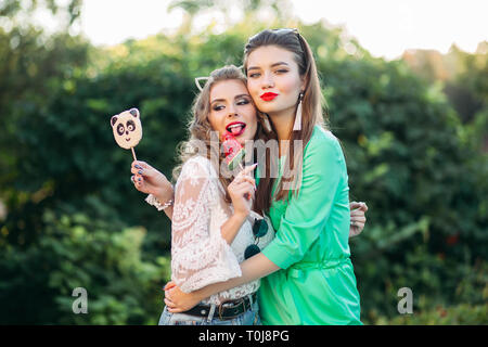 Two pretty and fashionable girlfriends holding candies on stick. Stock Photo