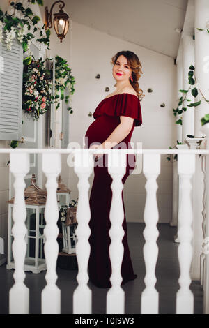 Charming pregnant woman in marsala dress posing on steps of house. Stock Photo