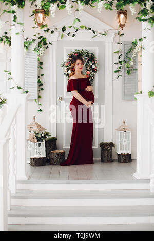 Charming pregnant woman in marsala dress posing on steps of house. Stock Photo