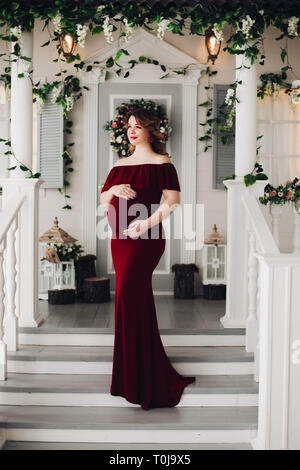 Charming pregnant woman in marsala dress posing on steps of house. Stock Photo
