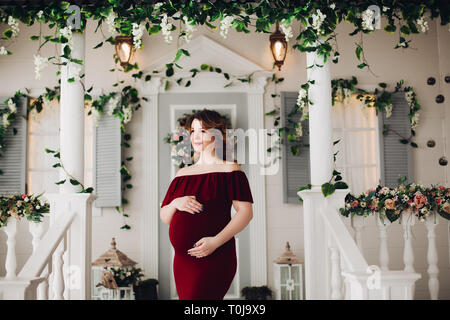Charming pregnant woman in marsala dress posing on steps of house. Stock Photo