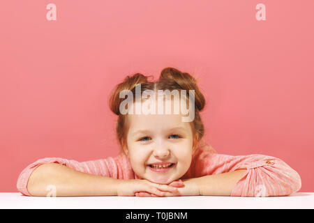 The little girl sits at the table, puts her head in her arms. Pink background. Copy space. Close-up. Stock Photo