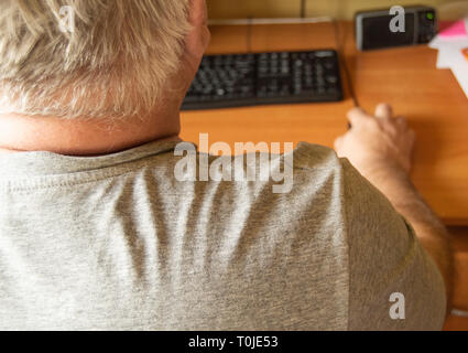 An elderly gray-haired man uses a computer mouse, work at home for the disabled, training pensioners to work on a PC, a view from the back. Stock Photo
