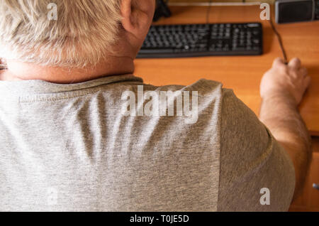 An elderly gray-haired man uses a computer mouse, work at home for the disabled, training pensioners to work on a PC, a view from the back. Stock Photo
