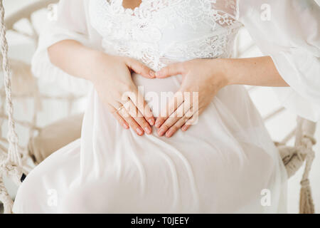 Beautiful blonde pregnant woman wearing in white long dress sitting at swing with flowers and looking, smiling at camera. Gorgeous mom expacting baby, Stock Photo