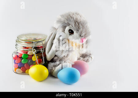 Stuffed toy easter bunny with colorful dyed eggs and a glass jar filled with jelly beans on a white background.  Copy space to the right. Stock Photo